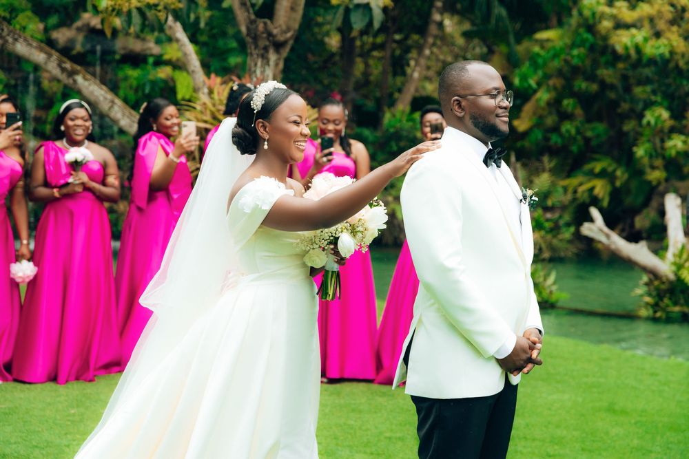 Bride in white gown and groom in white jacket celebrate with bridesmaids in bright pink dresses on garden lawn.