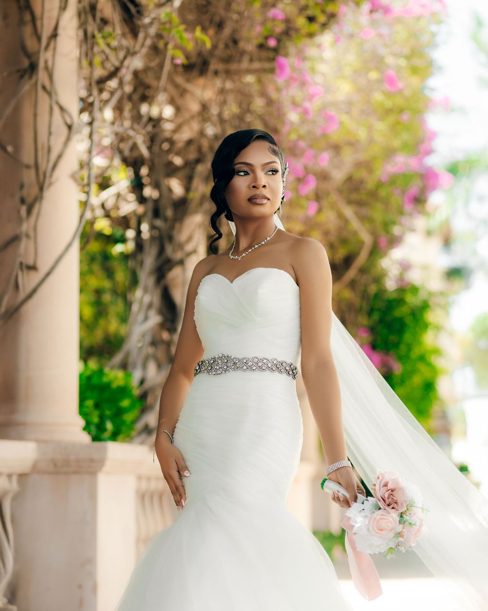 A stunning white mermaid wedding dress with beaded belt sparkles against a backdrop of pink flowers and stone columns.