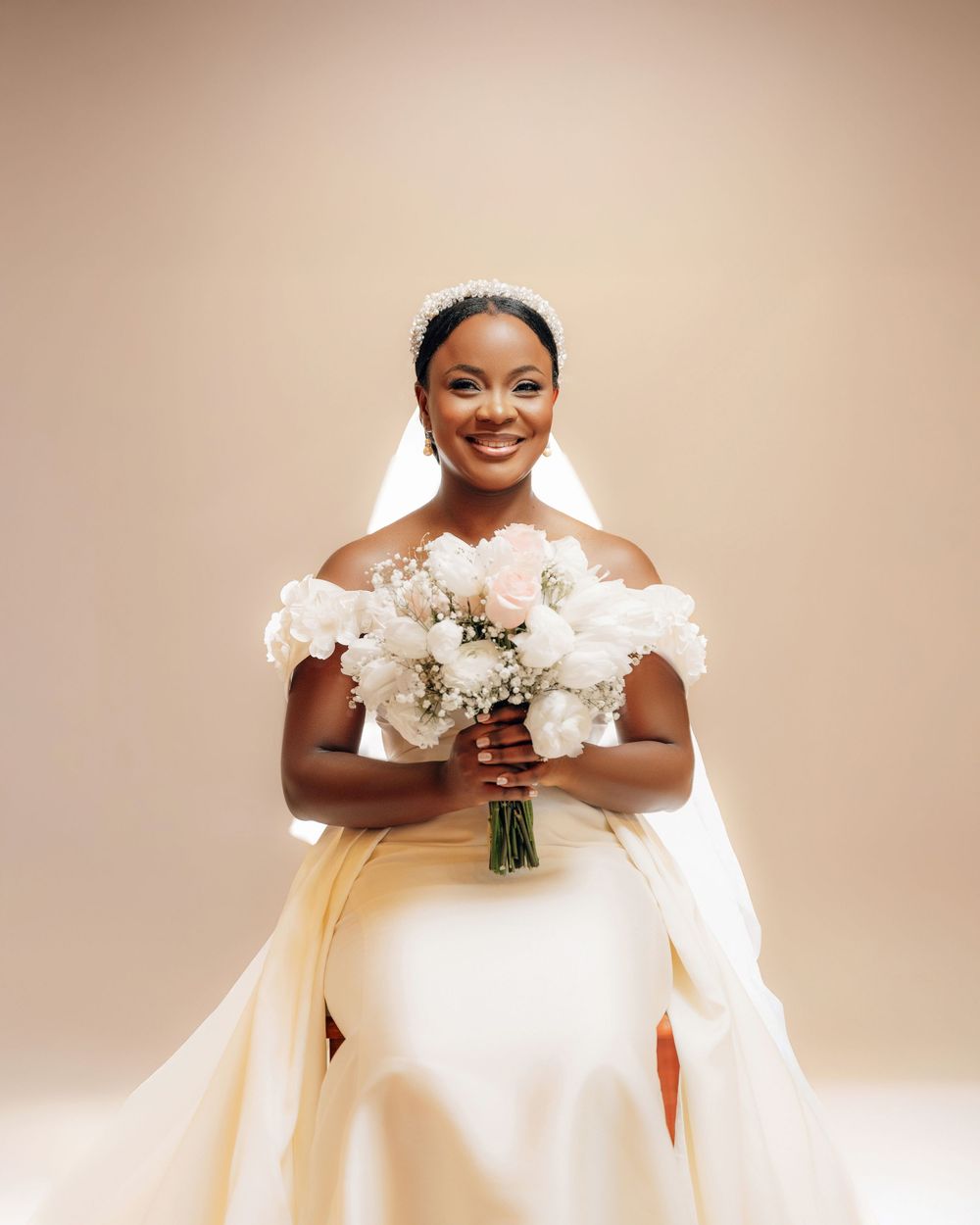 A joyful bride in a white off-shoulder wedding gown holding a cream and white bouquet against a beige backdrop.