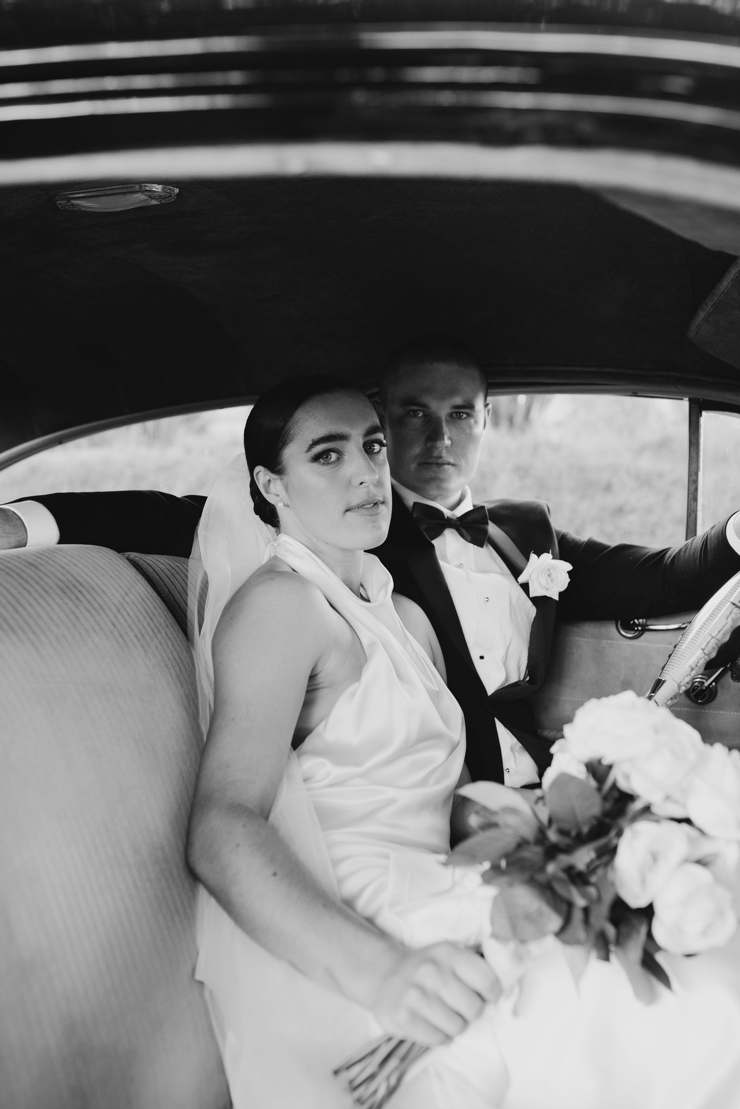 Bride and groom sit together in vintage car interior holding bouquet in black and white photo.