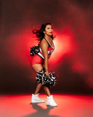 Portland Winterhawks Pro Dance Team Dancer in red and black uniform with pom poms poses dramatically against a red-lit background.