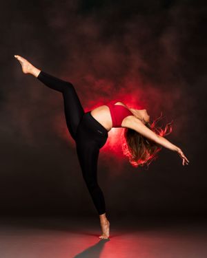 Portland Winterhawks Pro Dancer in a red top and black leggings performs graceful high kick against dark red backdrop with smoky lighting.