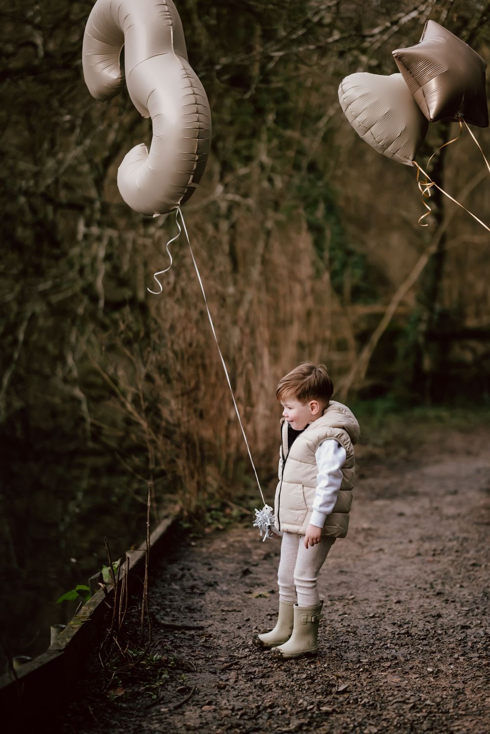 Child in white puffy jacket stands on forest path holding large white heart-shaped balloons.