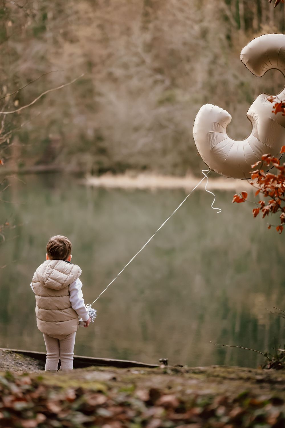 Small figure in puffy winter coat holds cream-colored balloon by lake on cloudy autumn day.