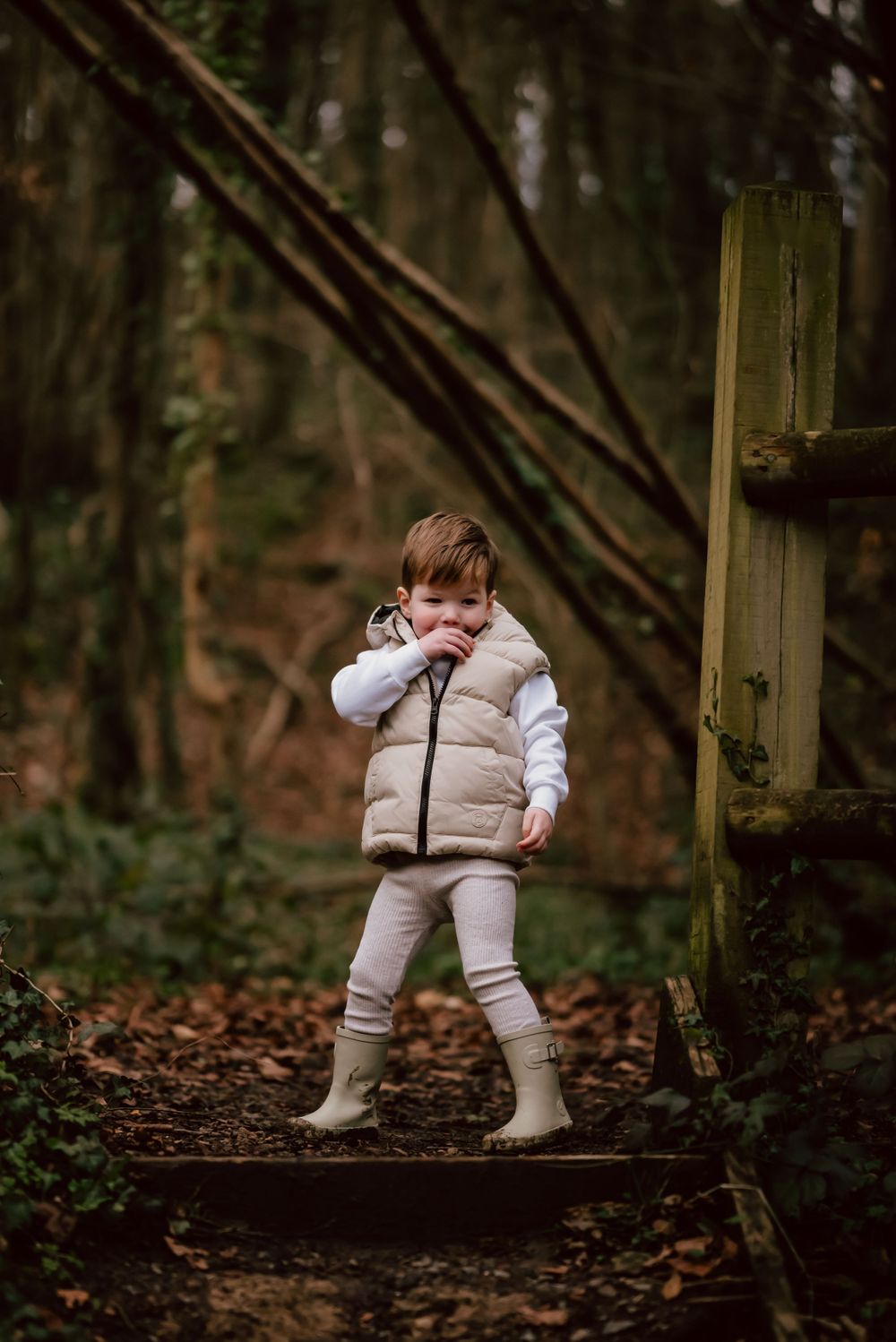 A small child in a white puffy vest and boots stands on a woodland trail during autumn.