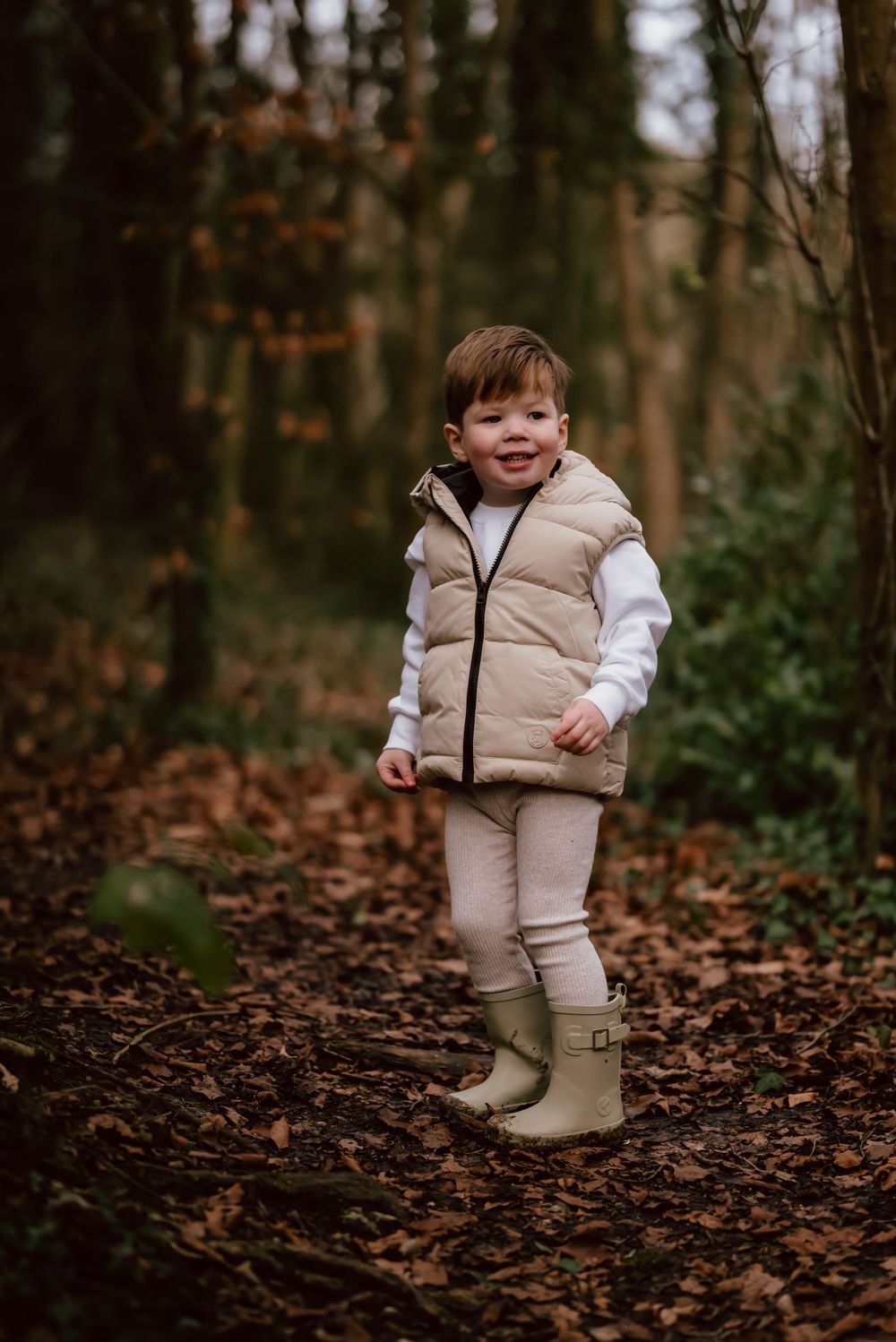 A small child in a beige puffer vest and rain boots stands on a leaf-covered forest trail during autumn.