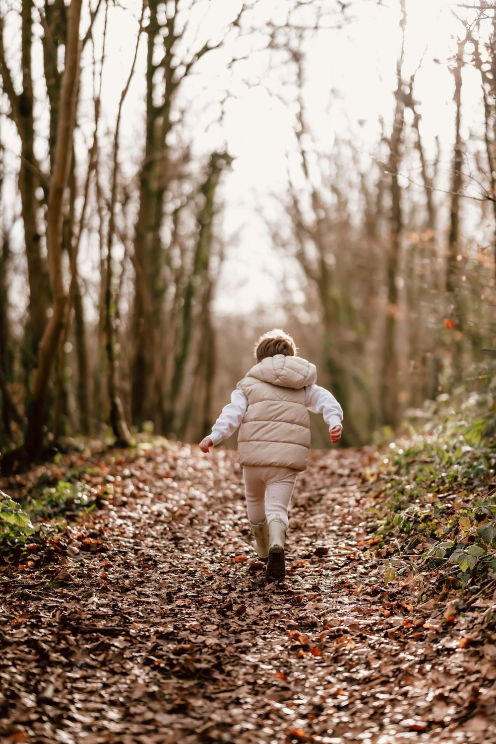 Small child in a puffy winter coat runs along a leaf-covered forest trail on an autumn day.