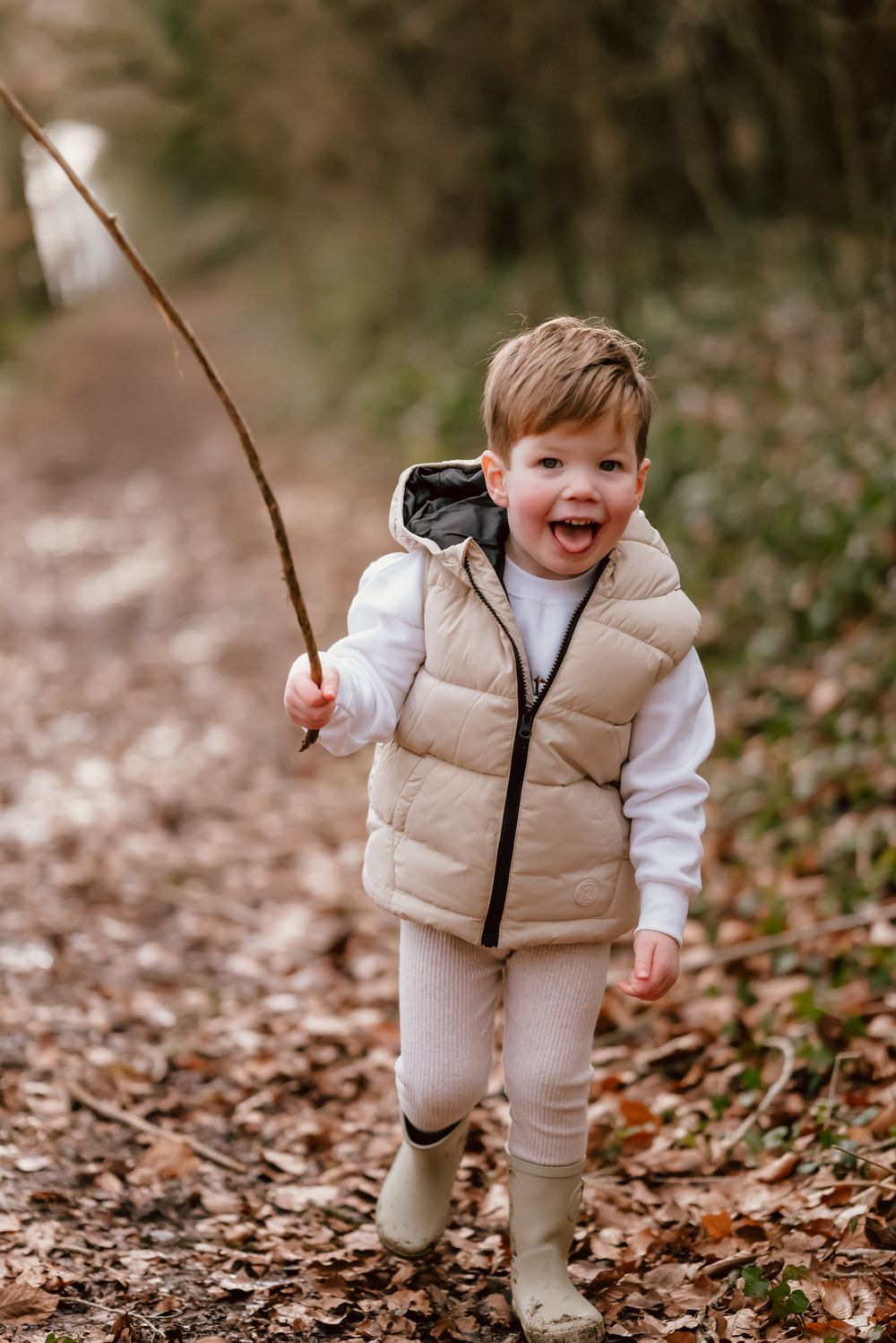 A young child wearing a beige puffer vest and gumboots walks along a forest path while holding a stick.