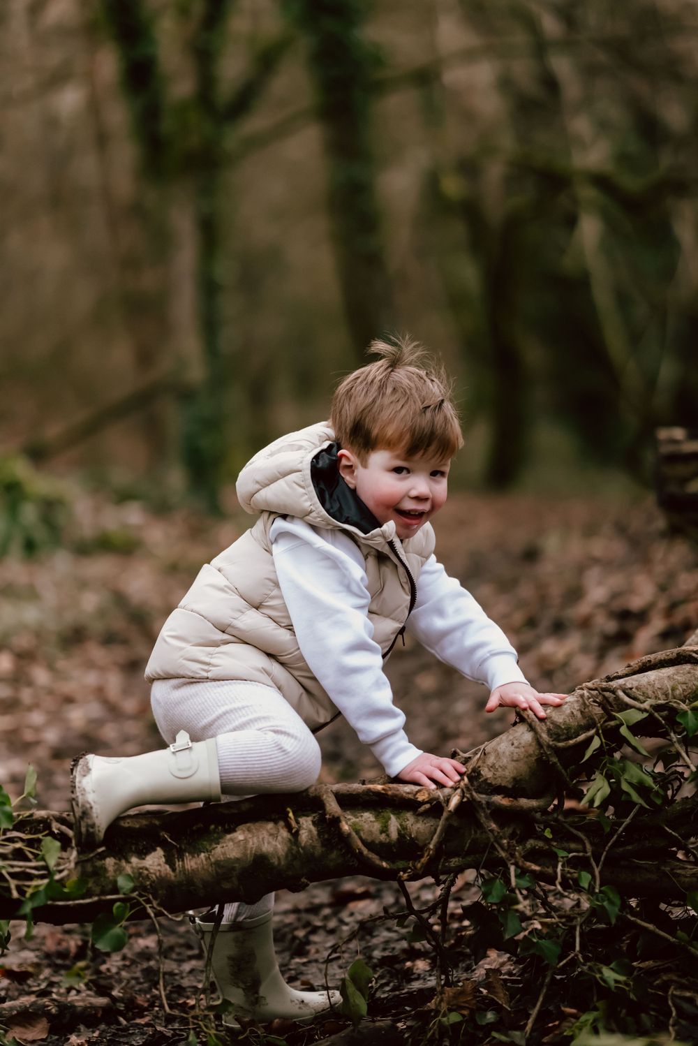 A small child in a cream puffer vest and white boots explores a fallen log in an autumn forest.