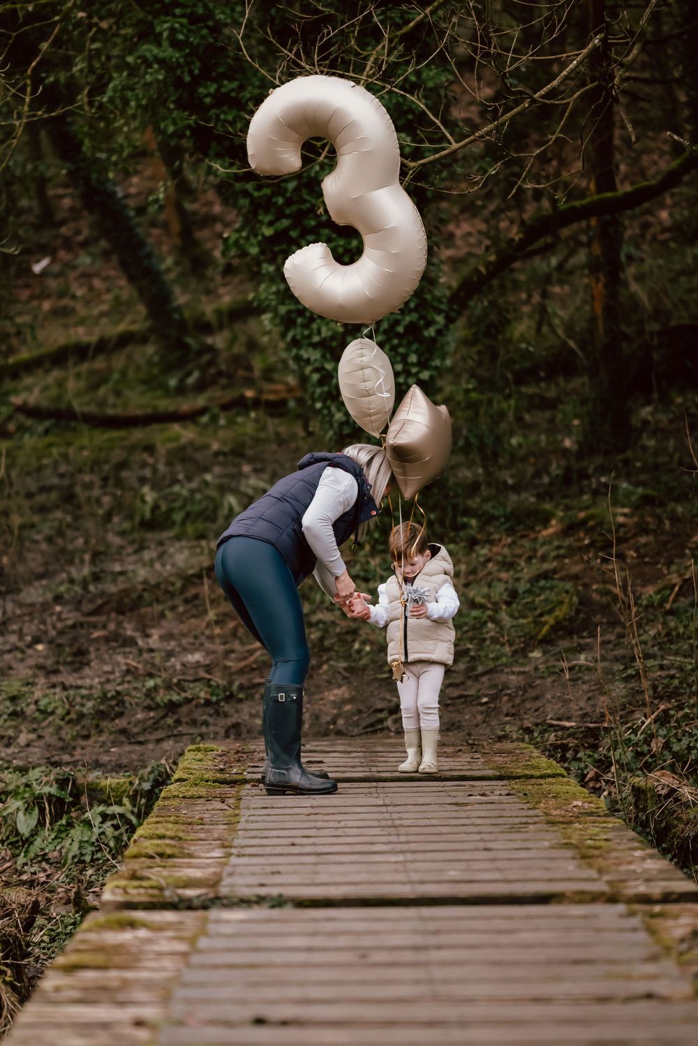 Two figures stand on a wooden boardwalk path in the woods with large white number 3 balloon floating above them.