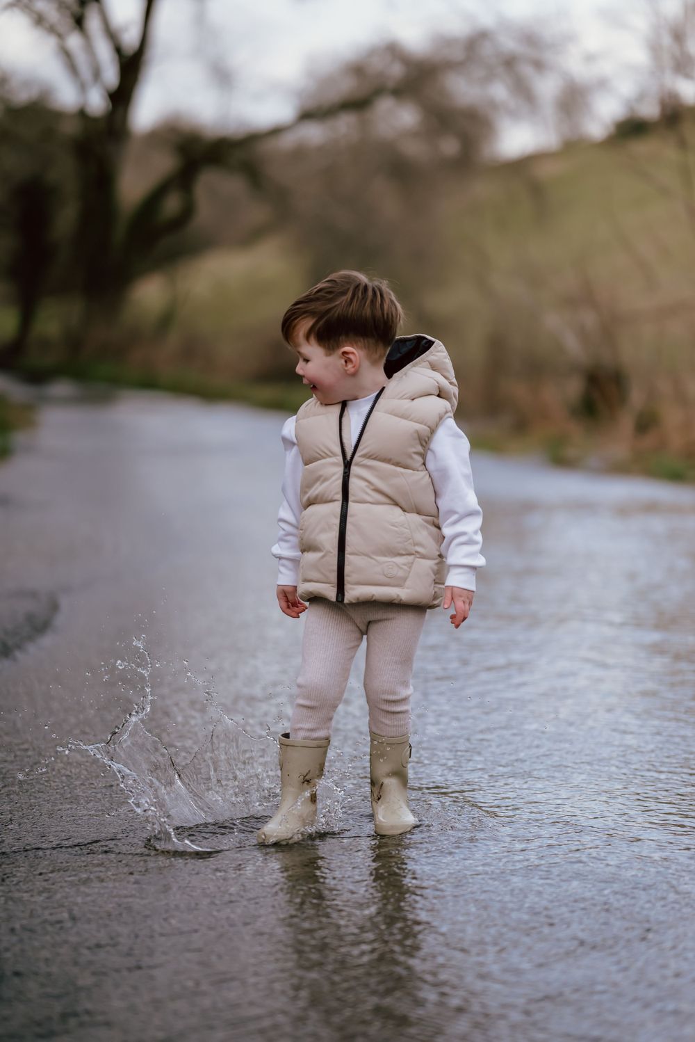 Young child in beige puffer vest and rain boots splashes in puddle on wet rural road during overcast day.