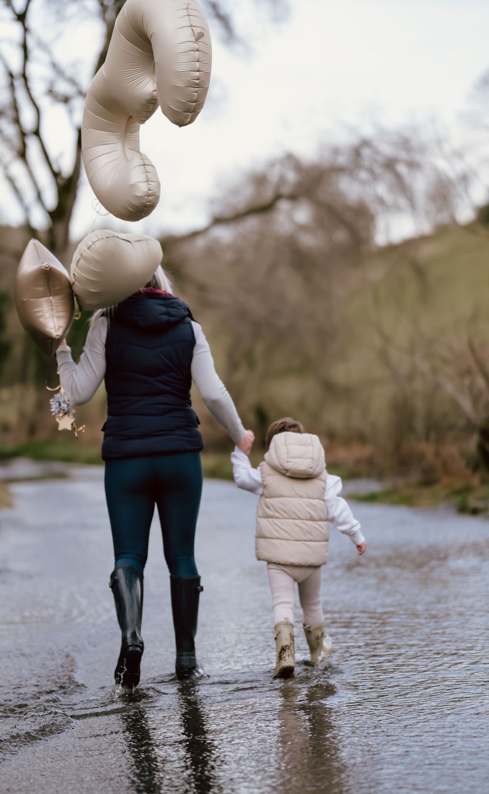 Two figures walk through puddles on a rainy day holding white balloons, wearing winter coats and rain boots.