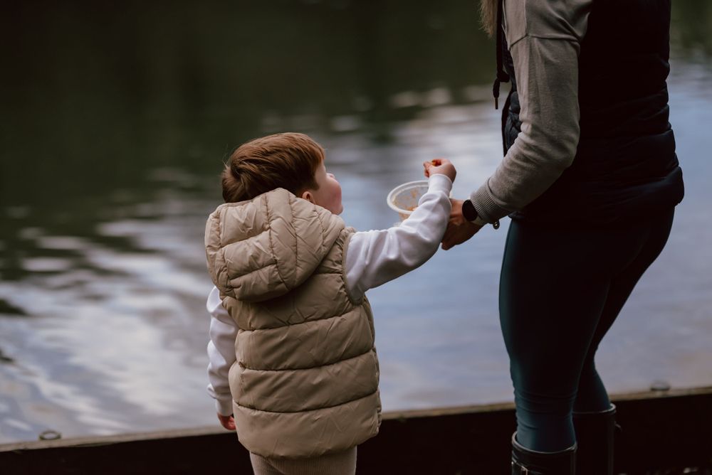 A small child in a puffy winter coat feeds the ducks with an adult while looking out at a peaceful lake.
