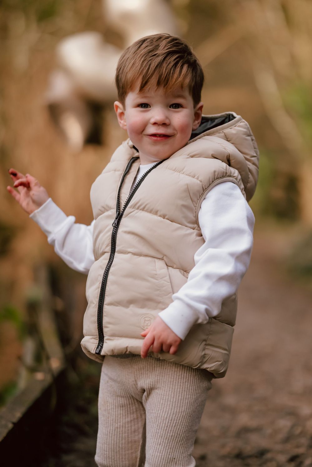 A small child wearing a beige puffer vest and white sweater smiles while standing outdoors in a natural setting.