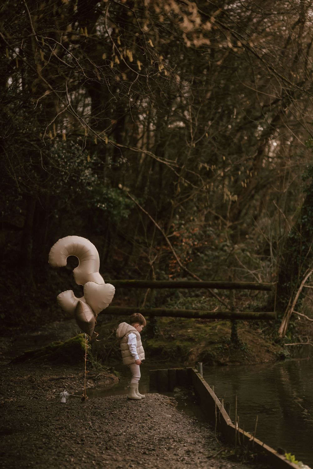 A small figure stands by a pond holding beige birthday balloons in a moody forest setting at dusk.