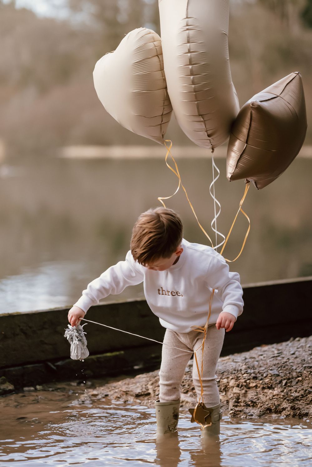 A small child in white clothing stands in shallow water holding balloons while playing with a boat at the shore.