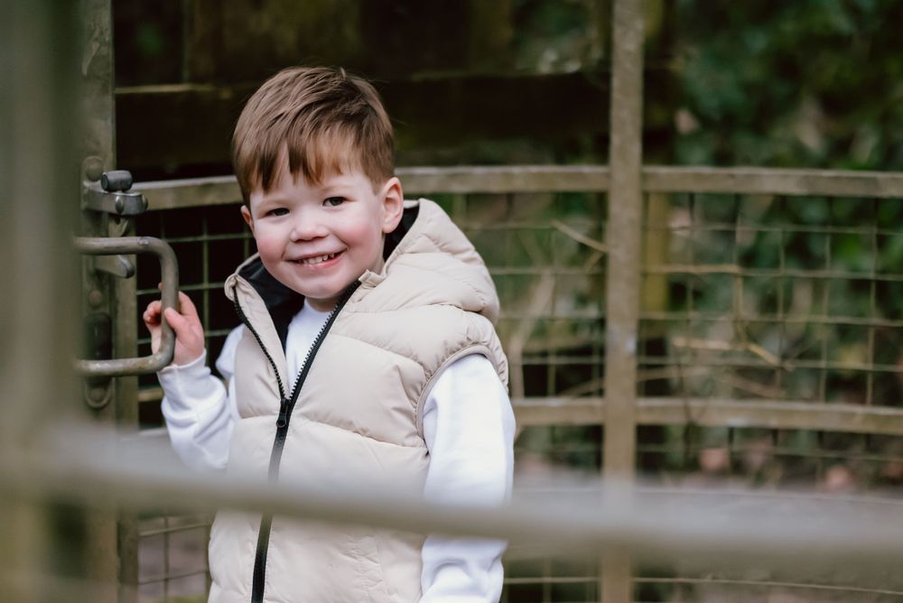 A young child wearing a puffy beige winter coat smiles while standing near a wooden fence outdoors.
