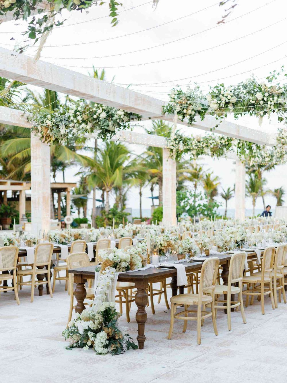 Elegant outdoor wedding reception setup with long wooden tables, white chairs, and floral arrangements under a draped canopy.