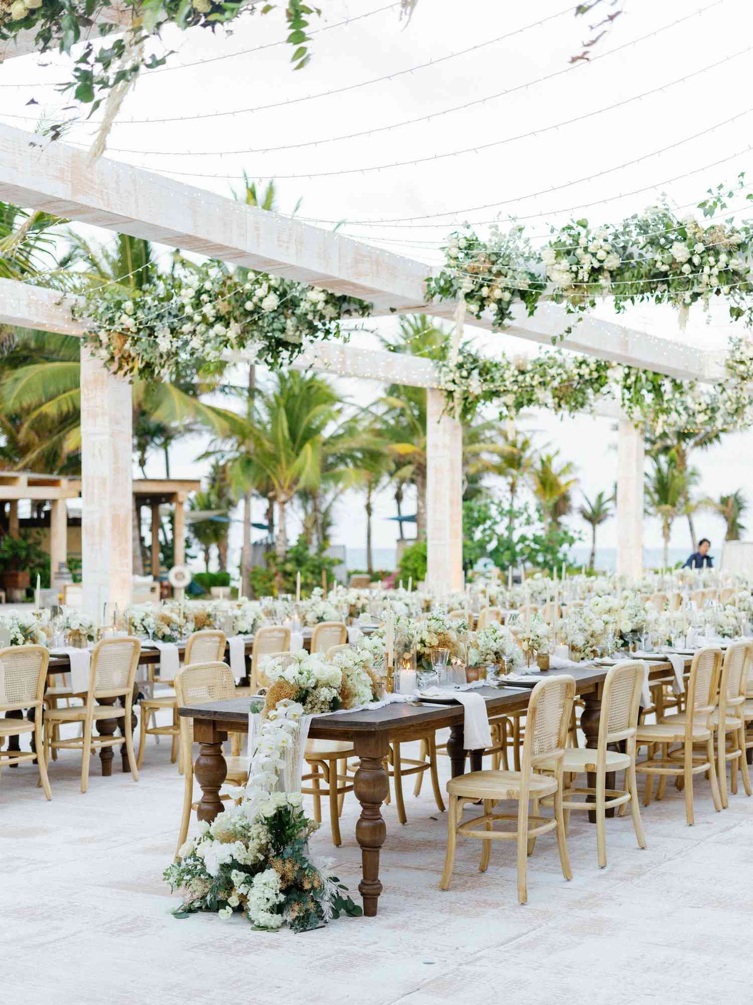 Elegant outdoor wedding reception setup with long wooden tables, white chairs, and floral arrangements under a draped canopy.
