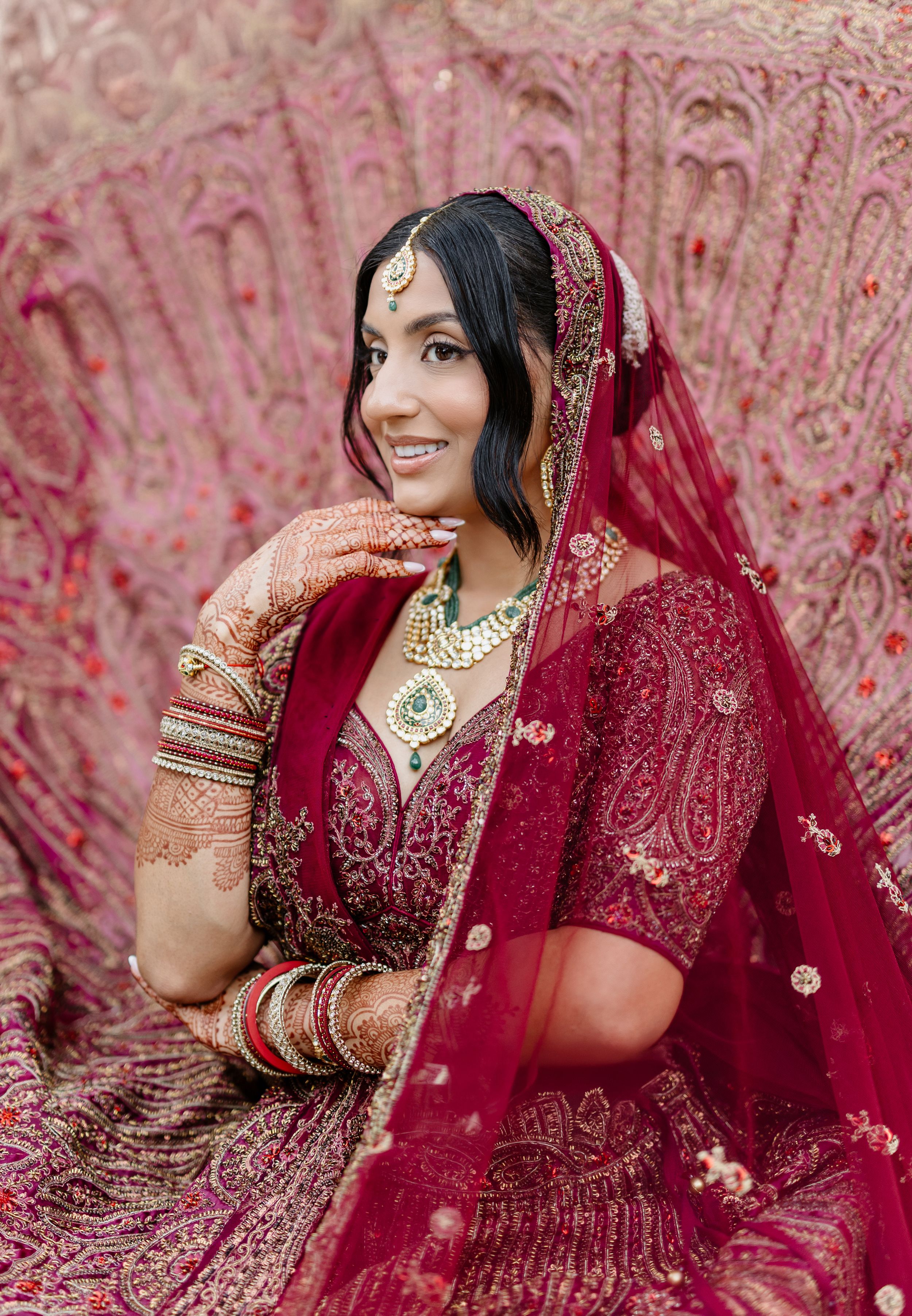 A bride in a traditional red Indian wedding lehenga poses elegantly against a pink floral backdrop.