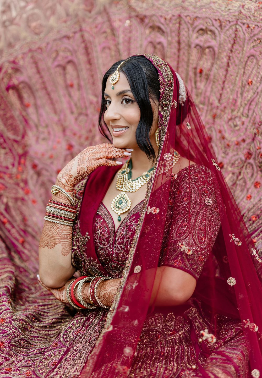A bride in a traditional red Indian wedding lehenga poses elegantly against a pink floral backdrop.