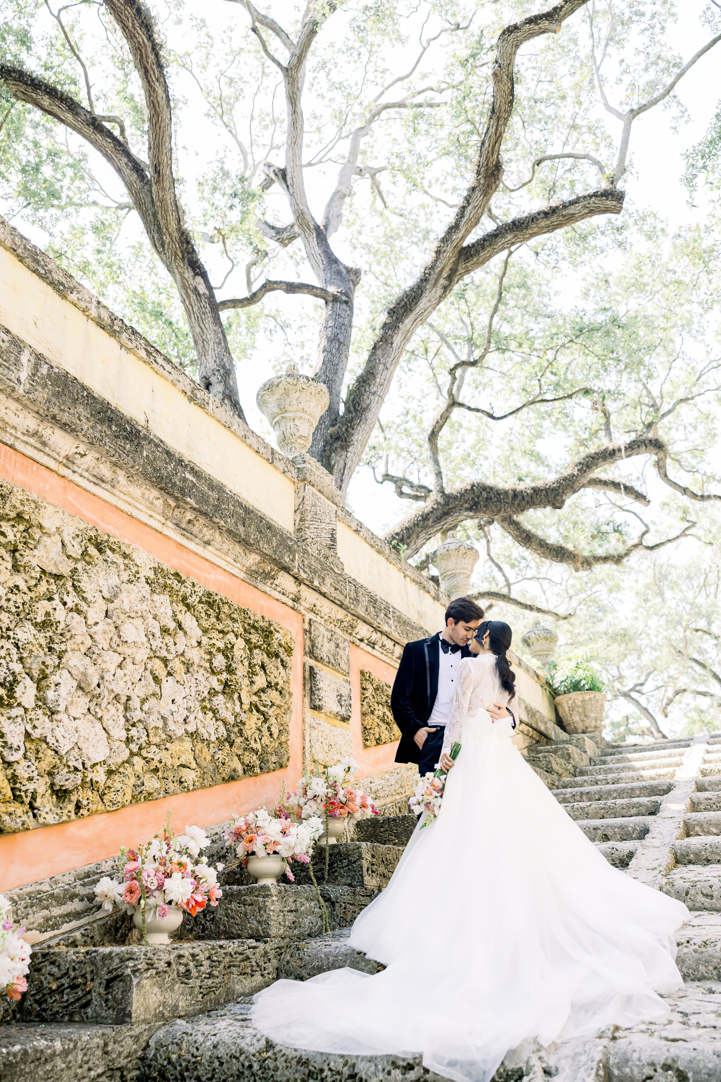 Elegant wedding couple embraces on stone steps beneath sprawling tree branches.