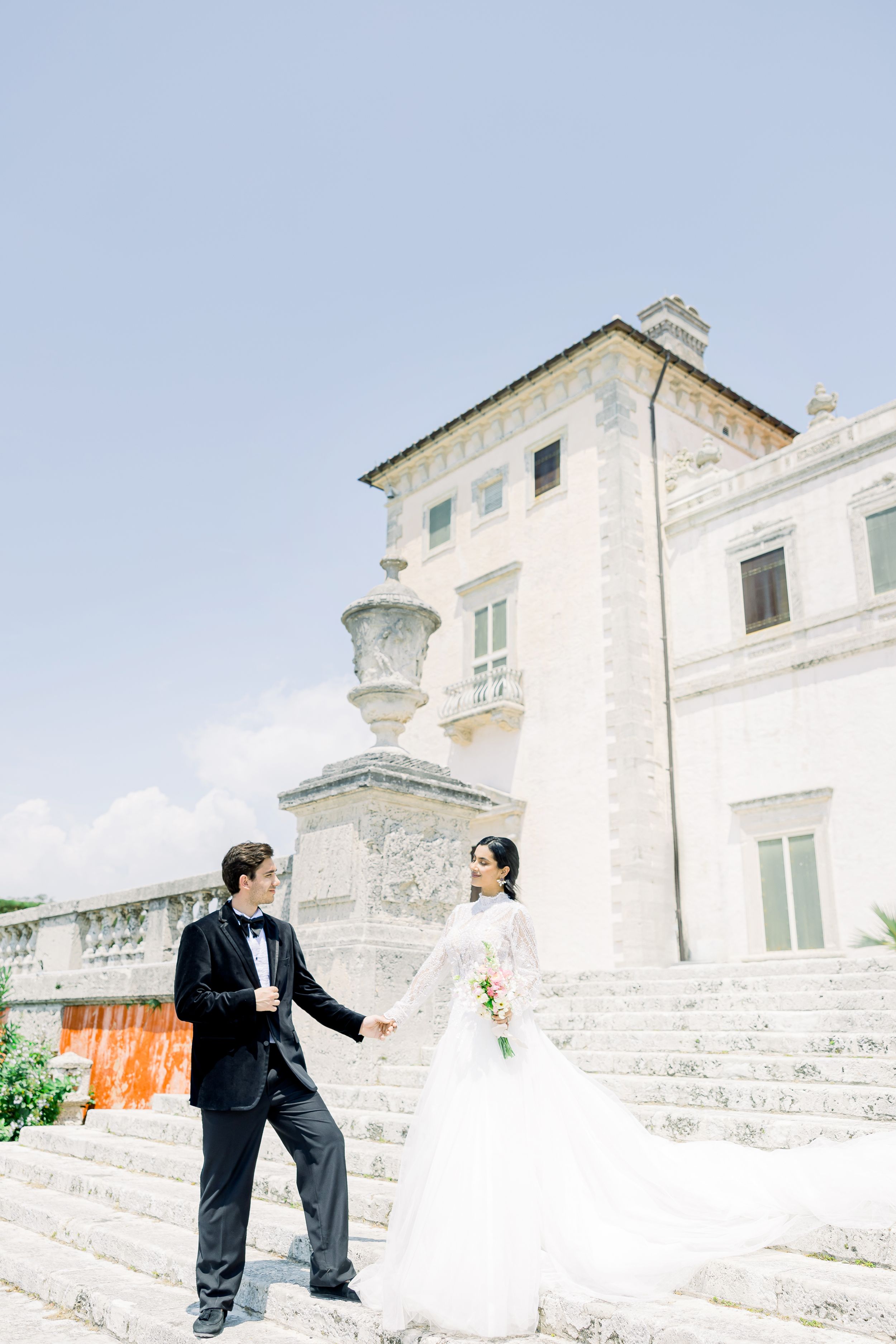 A romantic wedding photo of a couple on white stone steps against a classic Italian villa backdrop.