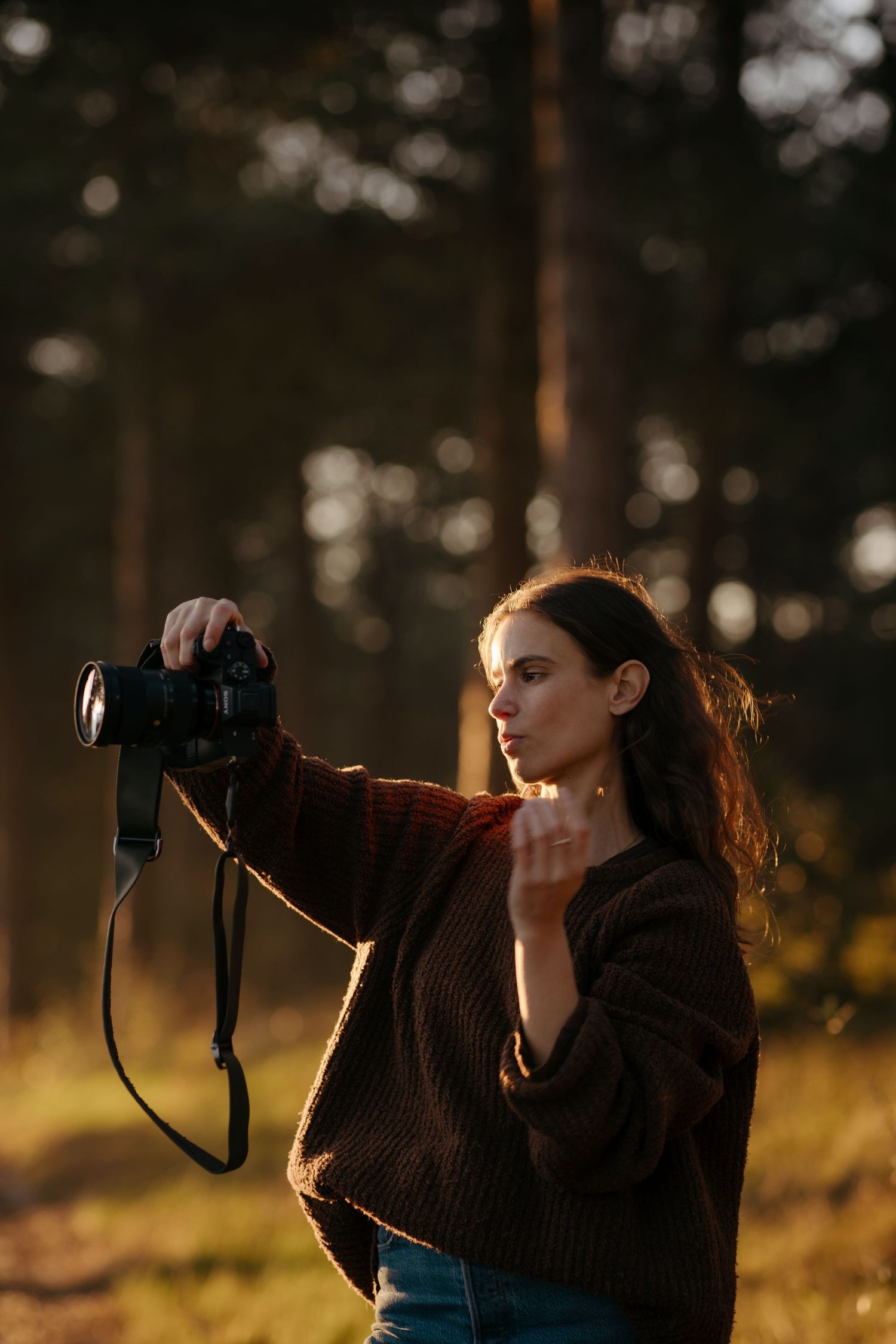 Photographer stands in golden sunlit forest while holding a camera during warm sunset light.