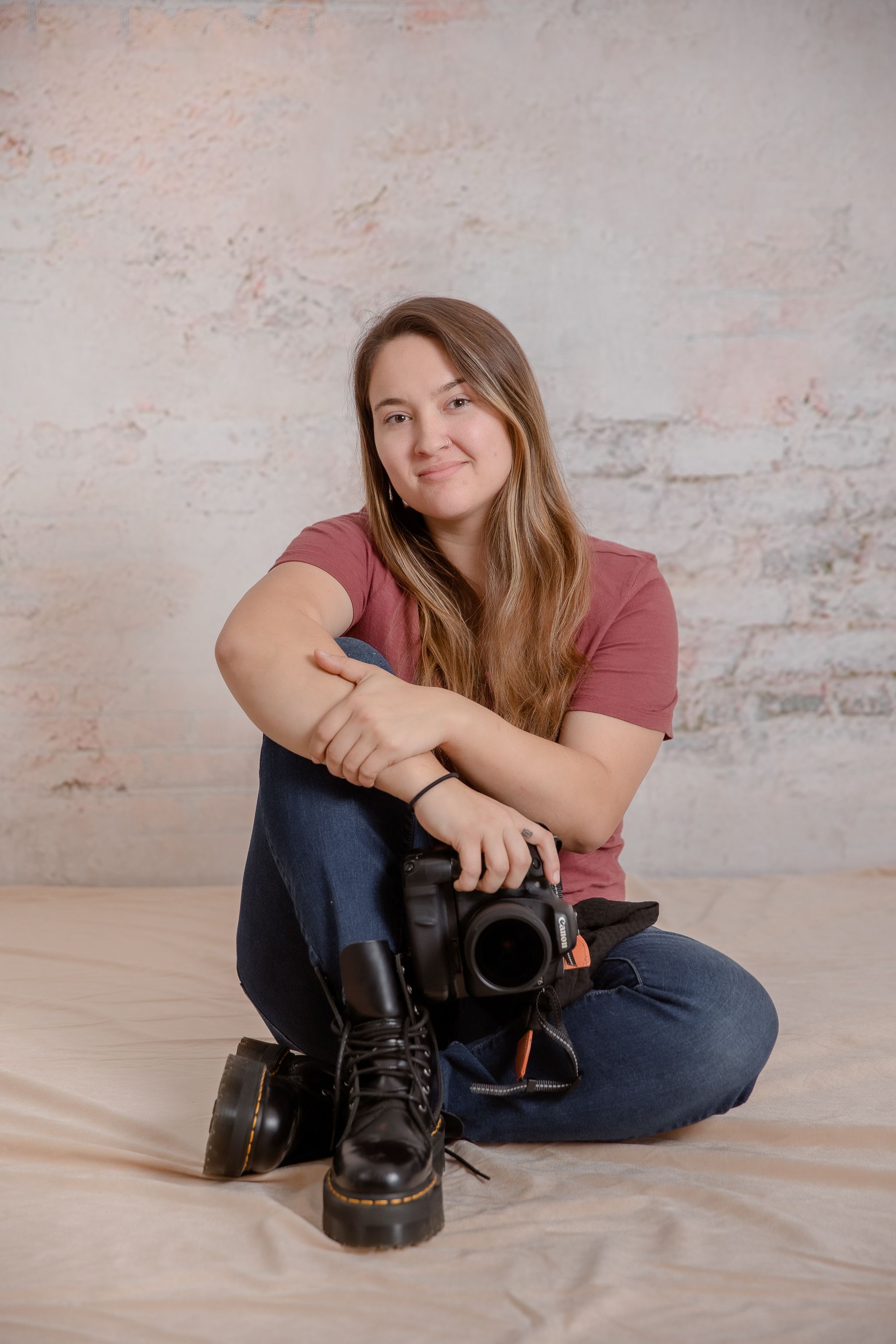 Portrait of photographer with camera sitting cross-legged against textured wall.
