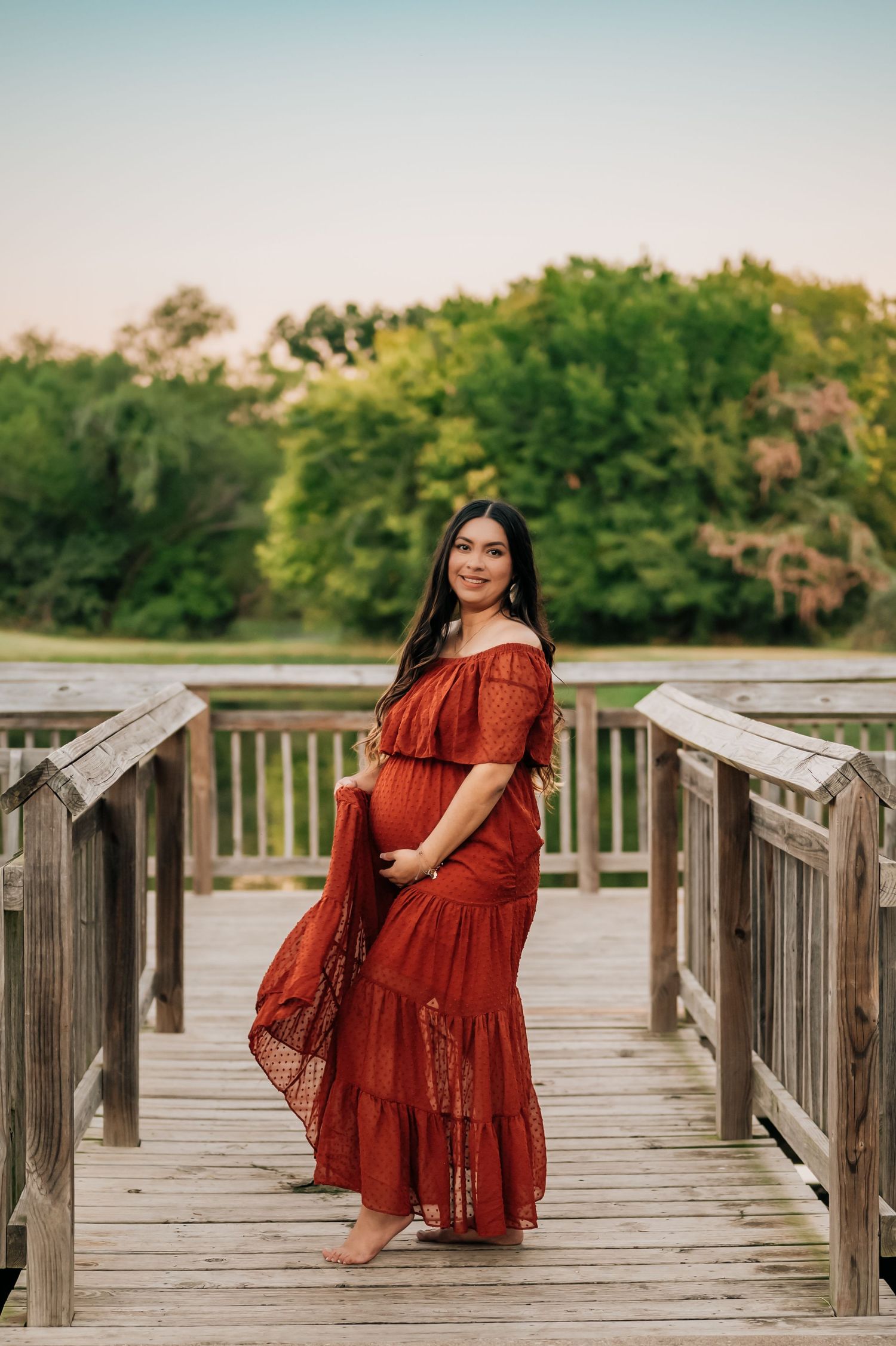 DFW MOM POSING WITH HAND ON BELLY DURING A MATERNITY PHOTOSHOOT IN COLLEYVILLE TEXAS. SHE IS STANDING ON A WOODEN DOCK AT SUNSET