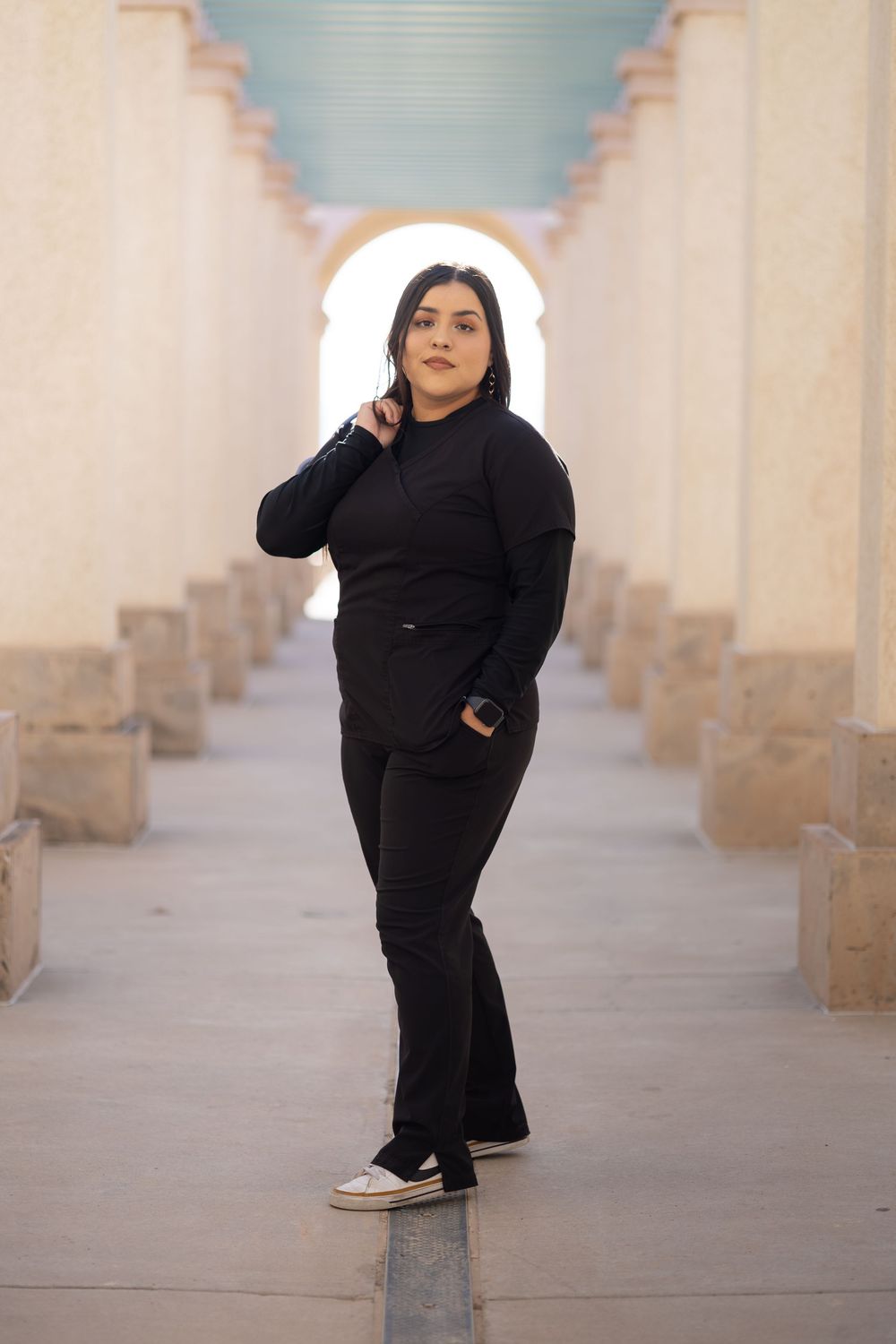 Young woman in all-black outfit posing in an arched corridor with stone columns and bright background.