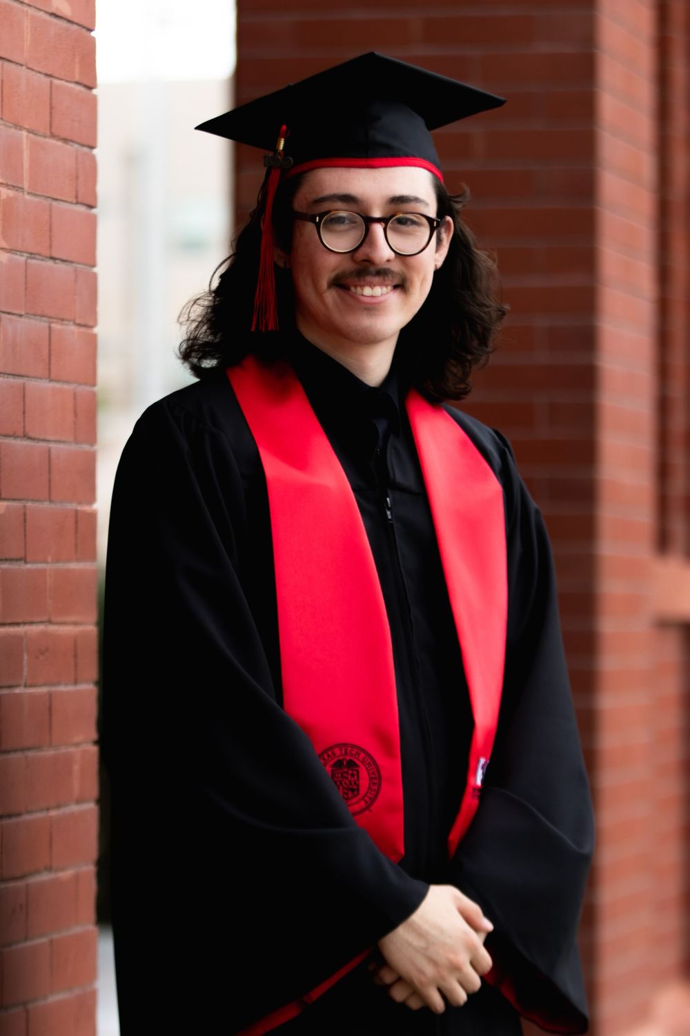 Graduate in black cap and gown with red sash smiles proudly near brick wall.