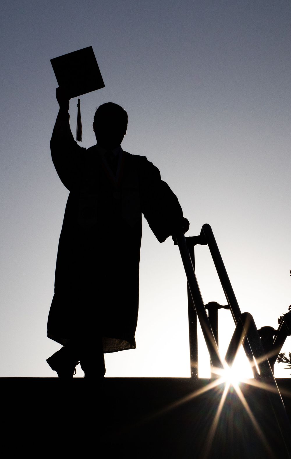 Silhouette of a graduate in cap and gown holding diploma aloft while using crutches, backlit by sunburst.