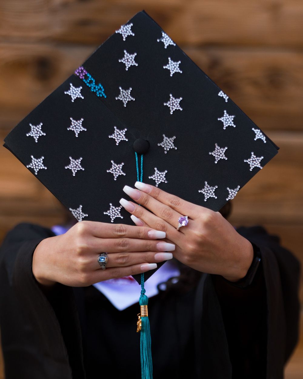 Person holding decorated black graduation cap with silver stars and teal tassel, wearing white nail polish and rings.