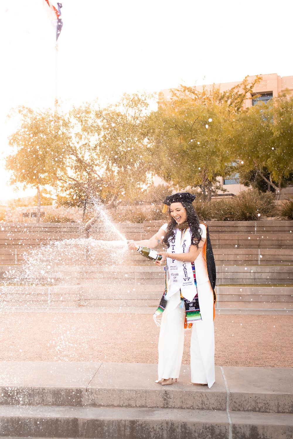 Graduate in white outfit and cap pops champagne bottle outdoors during golden hour celebration.