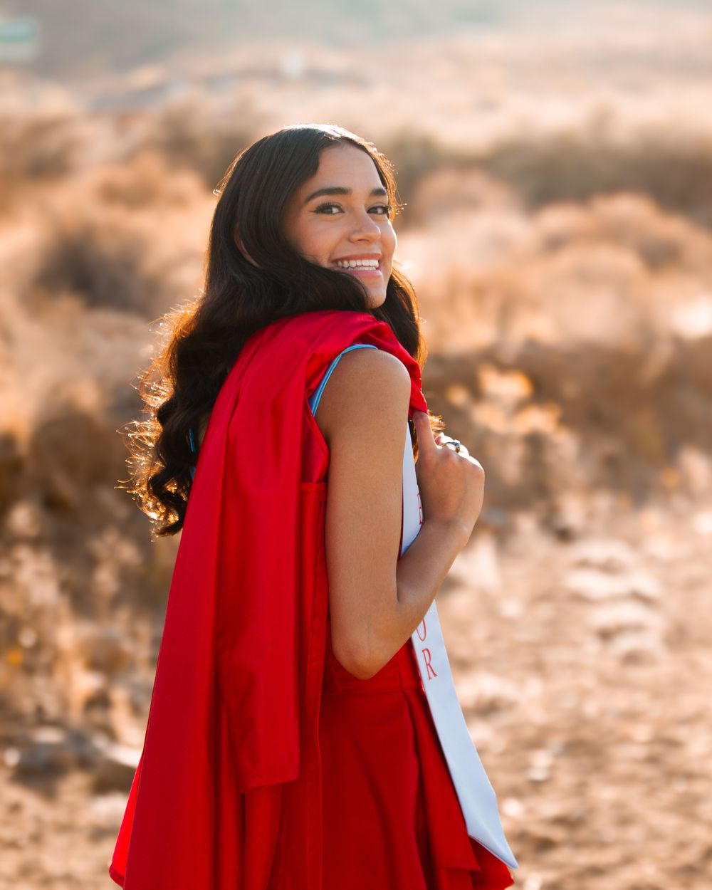 Young woman in red cape smiling over shoulder in golden field during sunset.