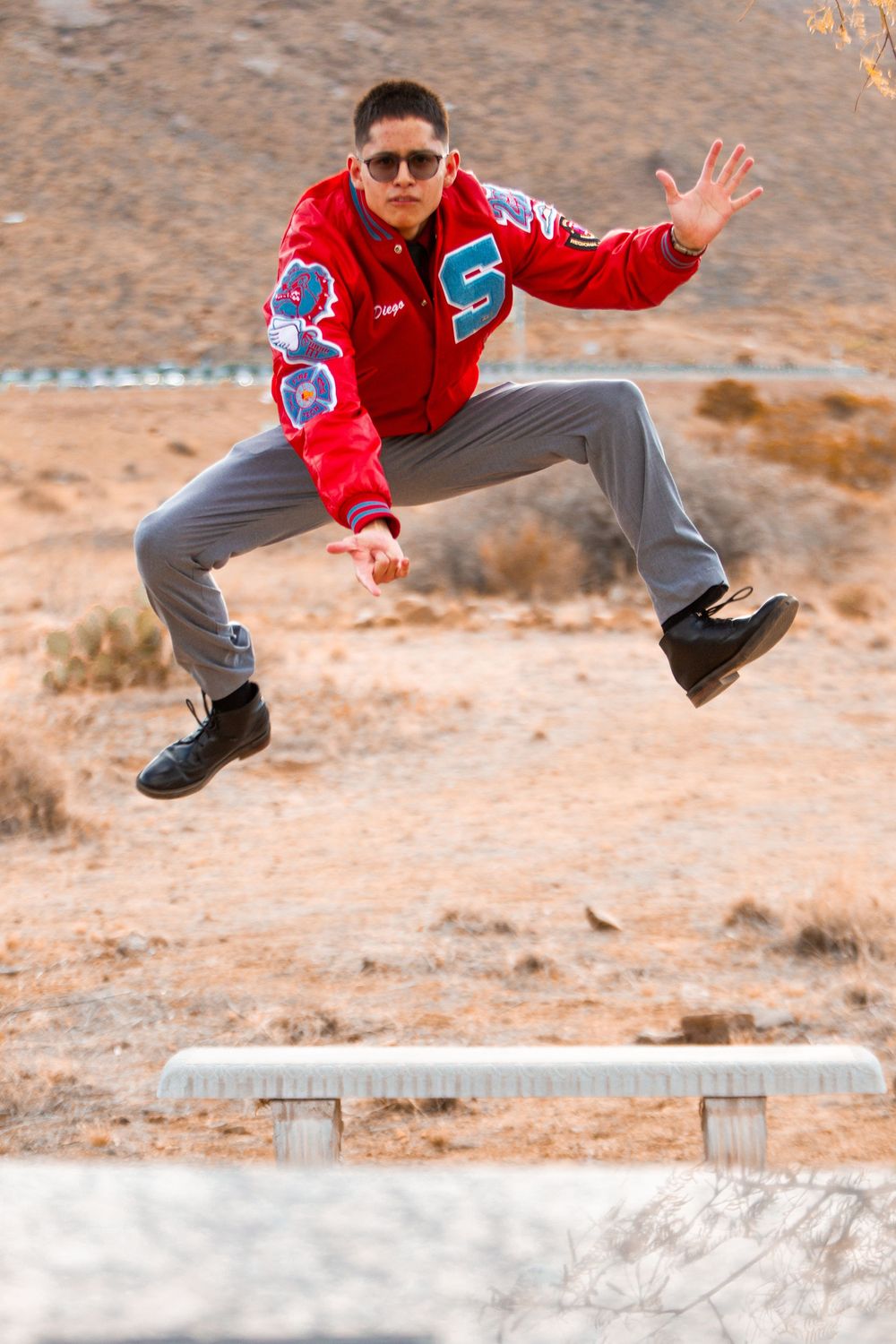 Young man in red varsity jacket jumping energetically over a bench in an arid desert landscape.