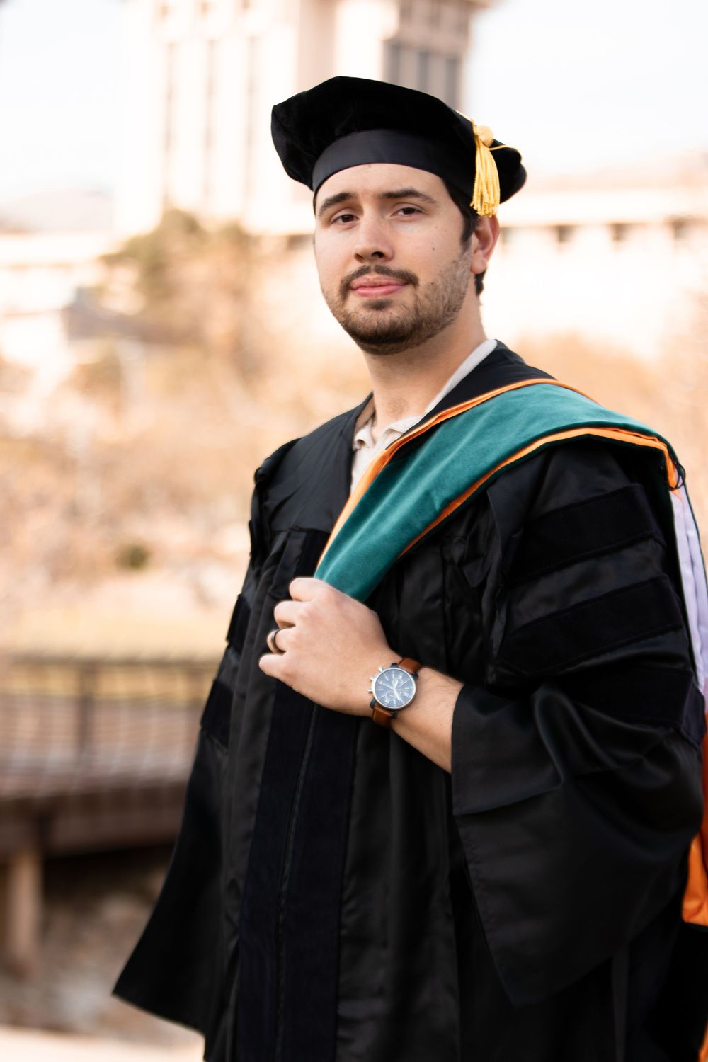 Young man in black doctoral graduation gown and cap with green and gold hood poses outdoors.