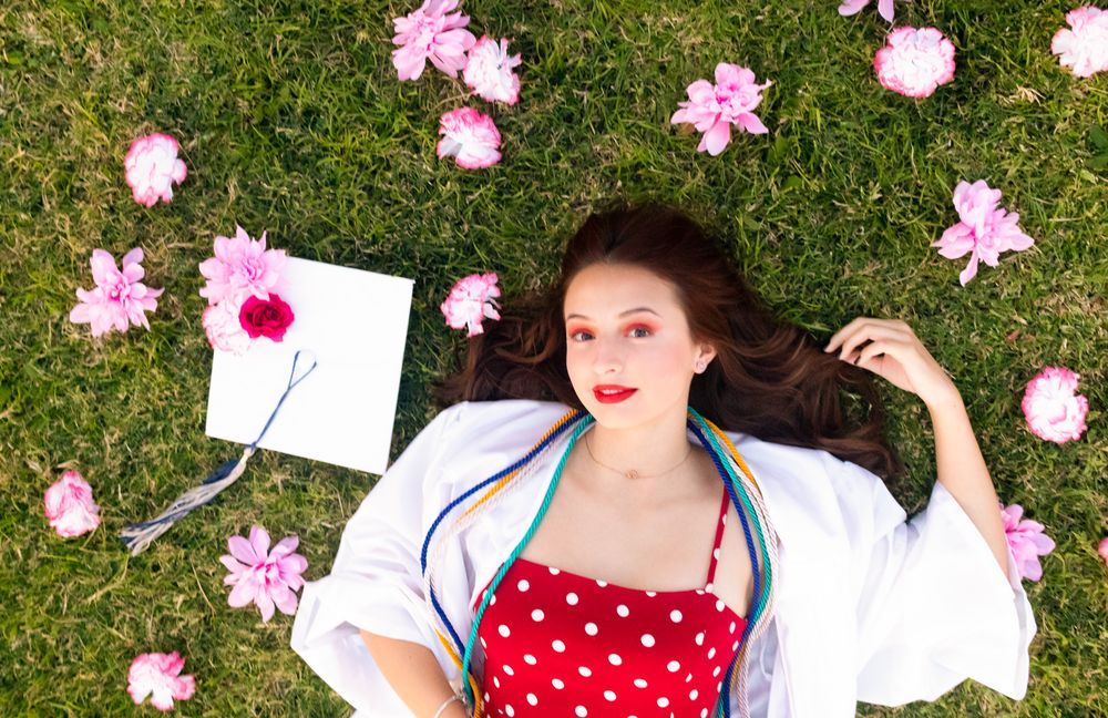 Woman in red polka dot dress lying on green grass surrounded by pink flowers and an open book.
