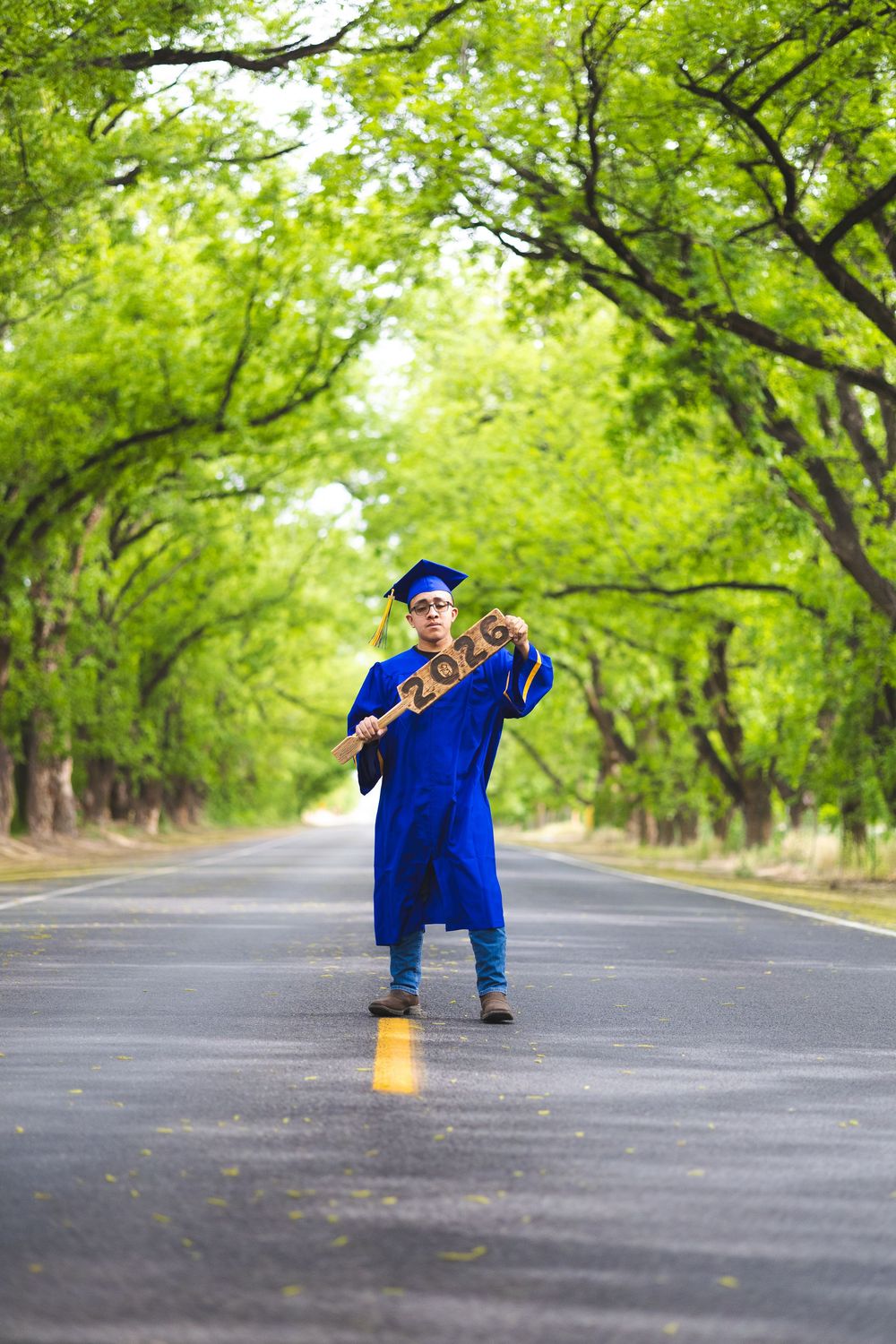 Graduate in blue cap and gown stands on tree-lined road holding 2023 wooden letters for graduation photo.