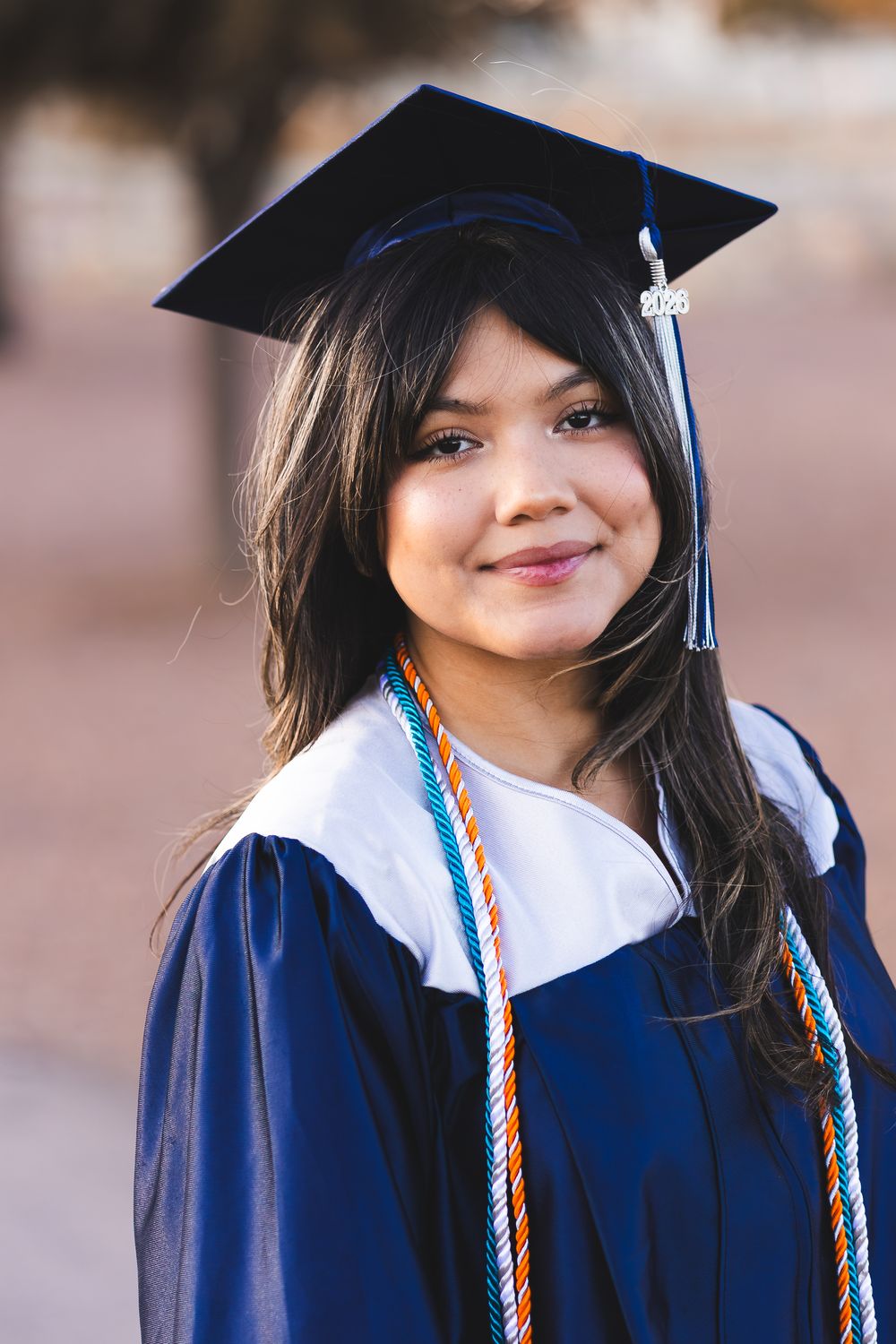 Young woman smiling in blue graduation cap and gown with honor cords outdoors.
