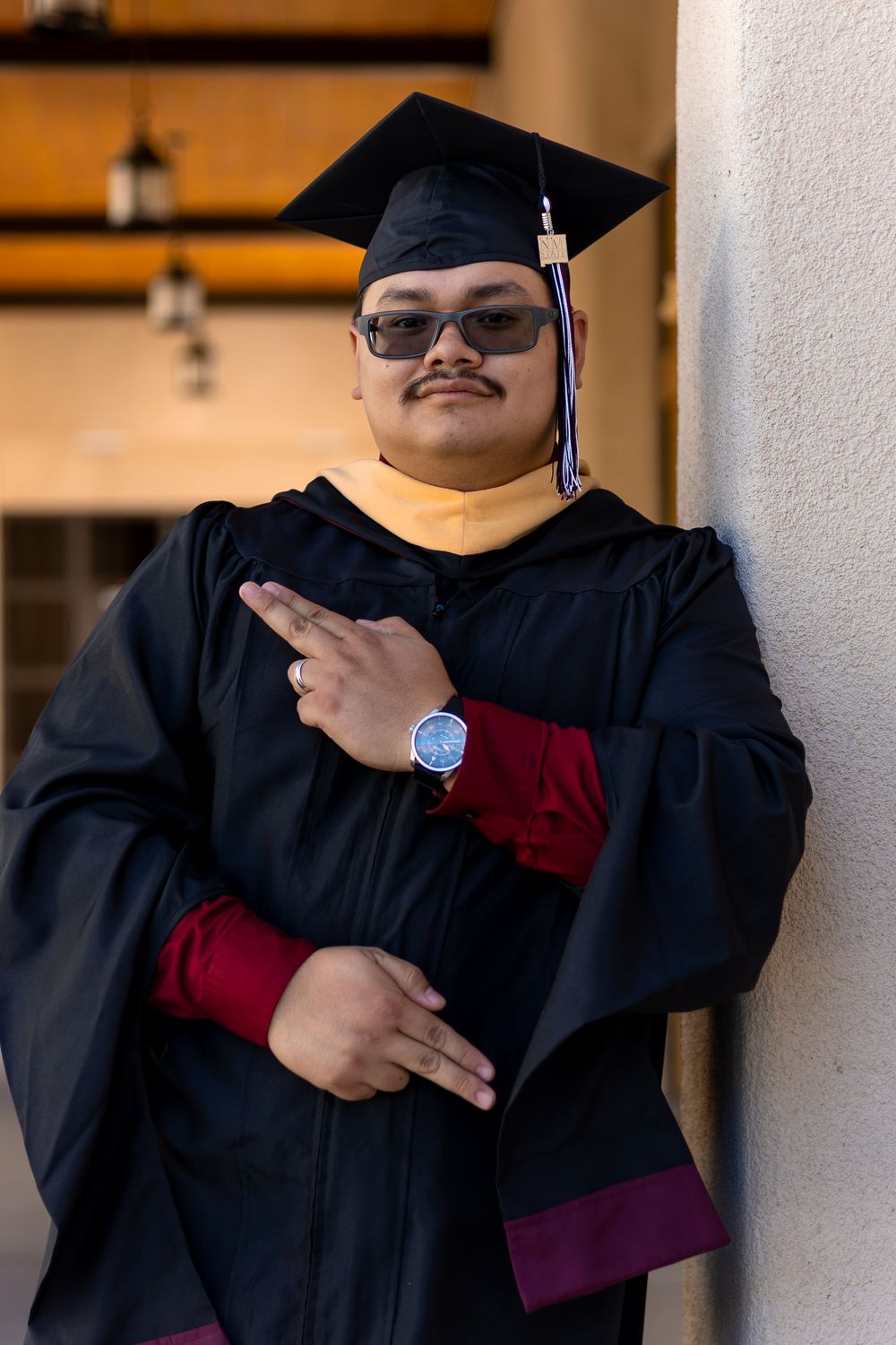 Graduate in black cap and gown with gold hood poses confidently, wearing sunglasses and a watch.