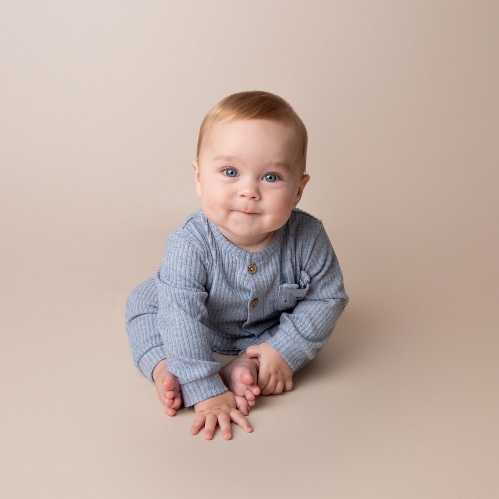 Smiling baby boy in blue striped romper sitting on a beige studio background.