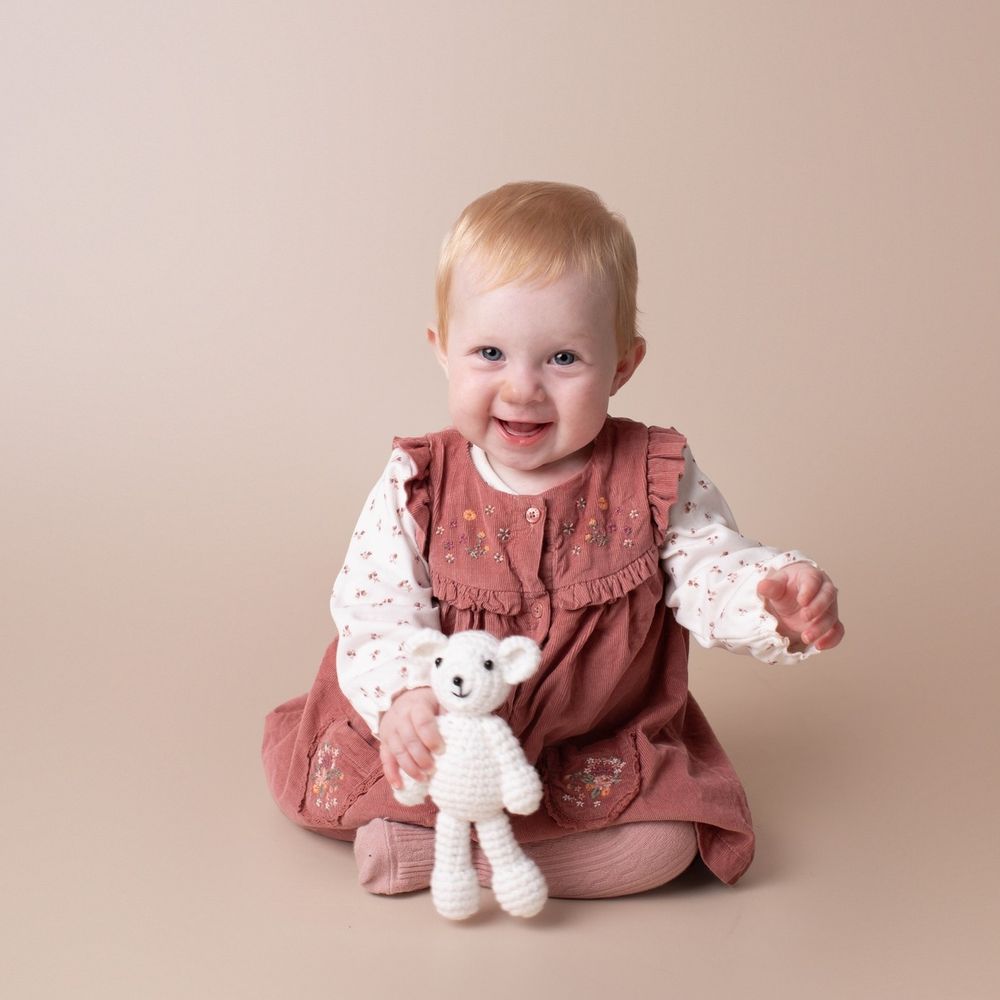 Happy baby girl in rose dress holding a white crochet teddy bear on beige background.