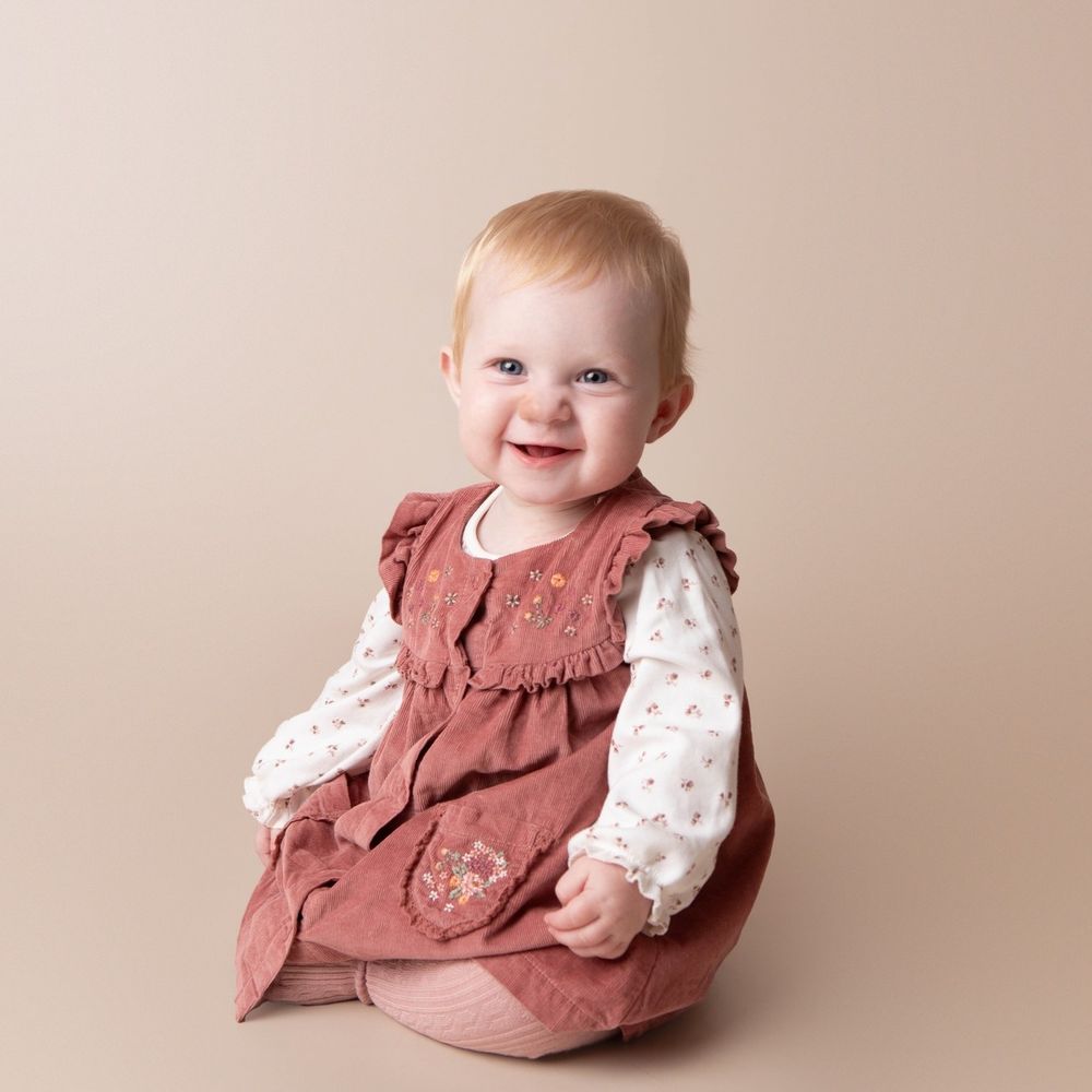 Cheerful baby girl in floral blouse and rose pinafore dress smiling on beige backdrop.