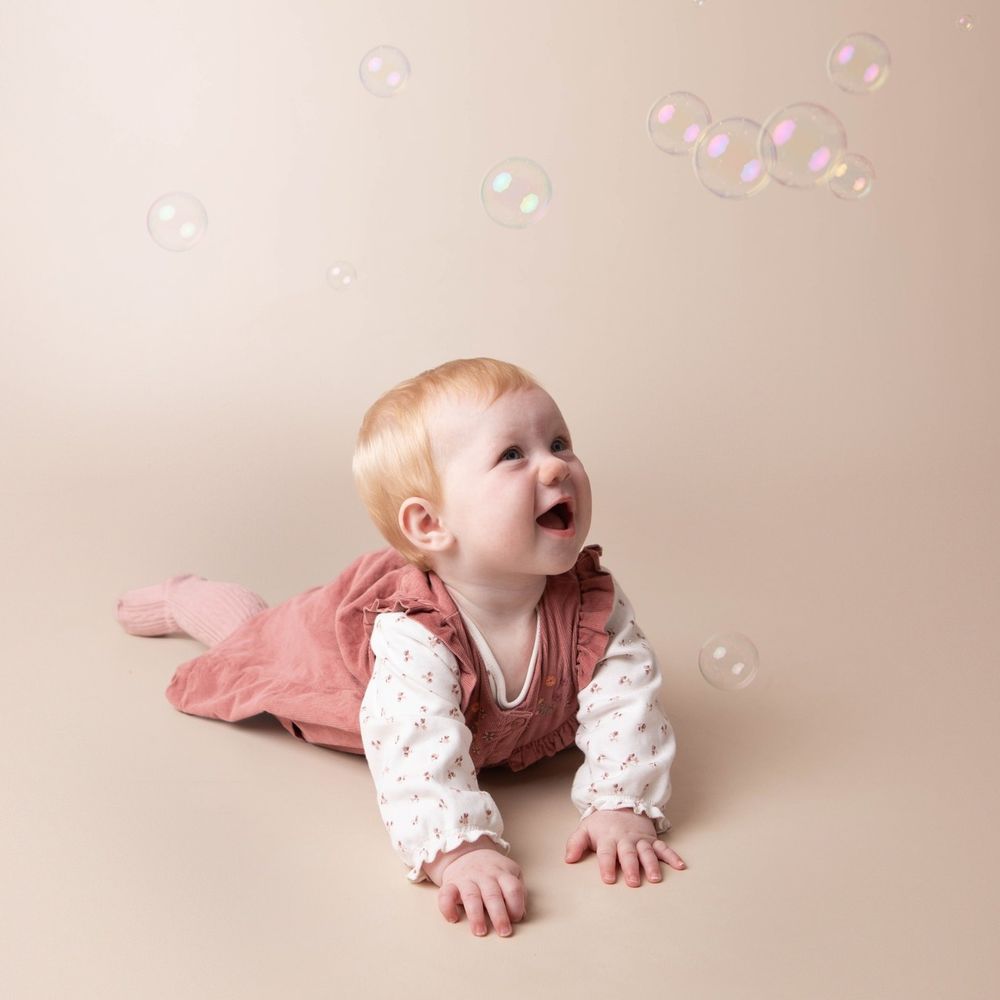 Laughing baby girl in rose dress doing tummy time while watching colorful bubbles float above.