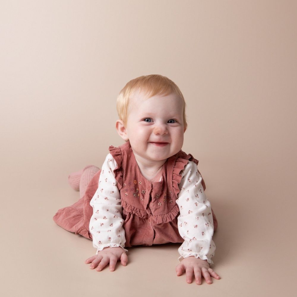 Smiling baby girl in rose pinafore and floral blouse crawling on a beige studio backdrop.