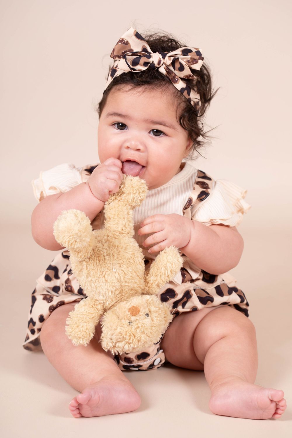 Baby girl in leopard print dress chewing on a stuffed bunny toy on beige background.