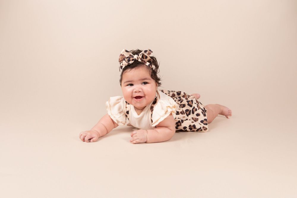 Smiling baby girl in leopard print outfit and bow headband doing tummy time on beige backdrop.