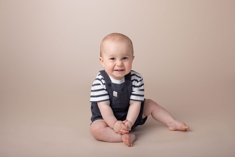 Smiling baby boy sitting on floor wearing striped shirt and navy overalls during studio portrait session.